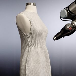 Still image of a white knit dress being formed to fit over a mannequin by a nearby robot arm hovering near it