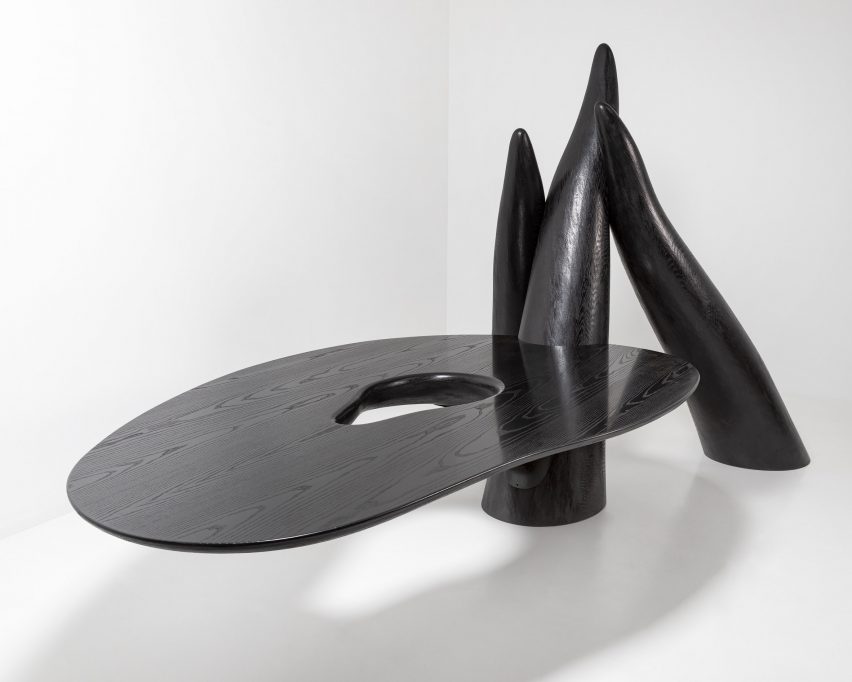 Photo of a black sculptural object with a large, tabletop-like surface extending from a collection of three, tentacle-like pillars rising from the floor