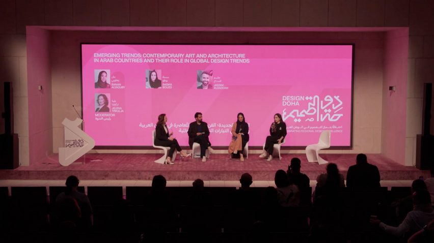 Video still of speakers on stage at the Design Doha Forum with a screen showing a bright pink graphic behind them