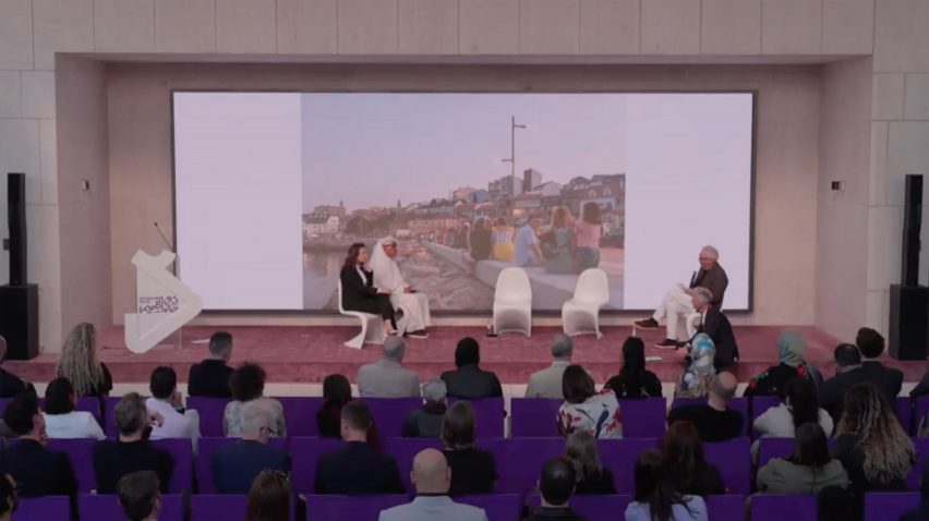 Still image of a speaker giving a presentation during a panel discussion, while an image of young people seated on a long stone bench on a seafront appears on the screen behind them