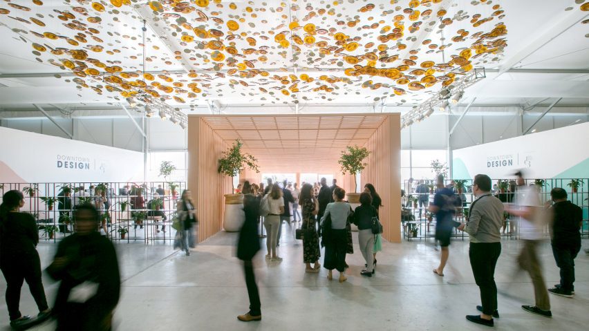 A photograph of a design fair, in tones of white, pink and orange, with multiple people in the space.