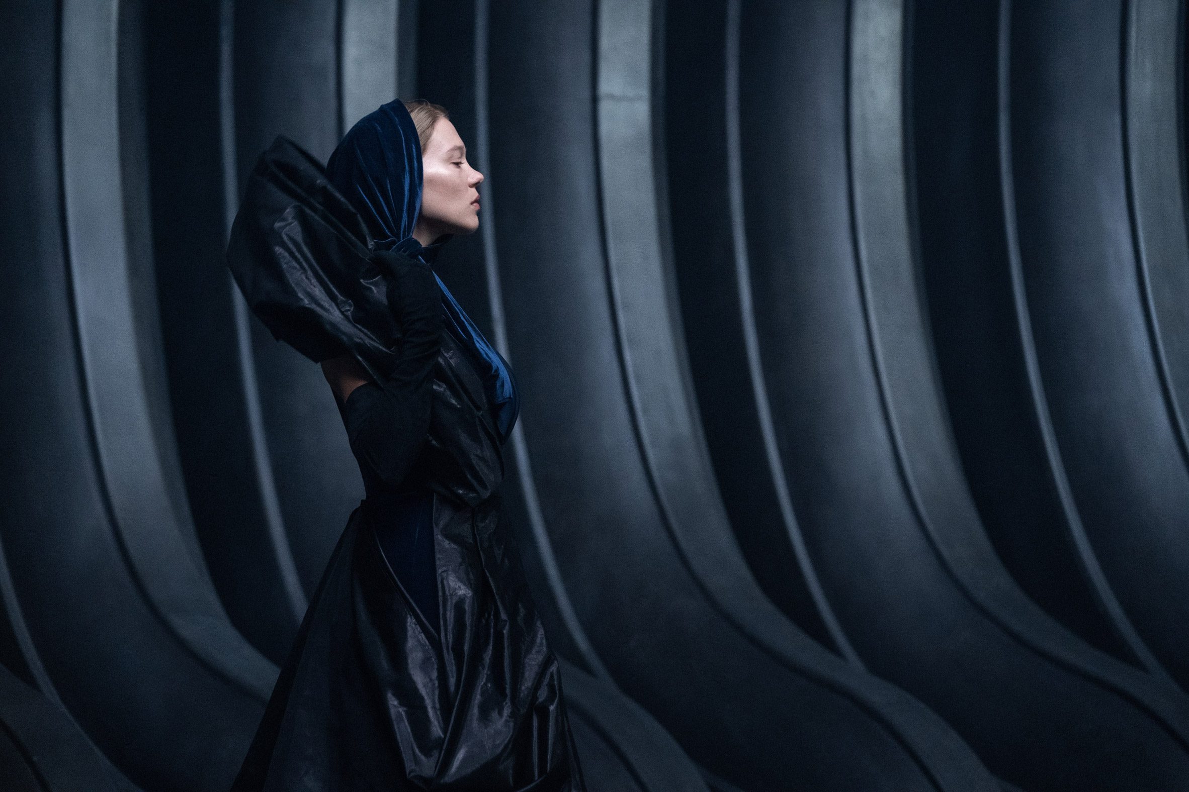 Dune Part Two production photo of Margot Fenring walking through the dark hallways of Giedi Prime against a background of ribbed, black walls that look made of moulded rubber