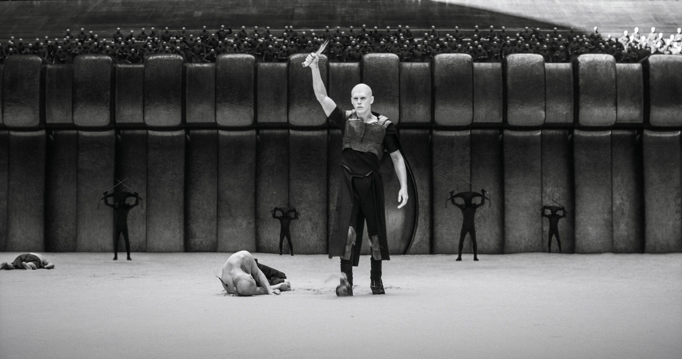 Dune Part Two production photo showing Feyd-Rautha Harkonnen holding his knife up in victory in a battle arena. The image is in black and white
