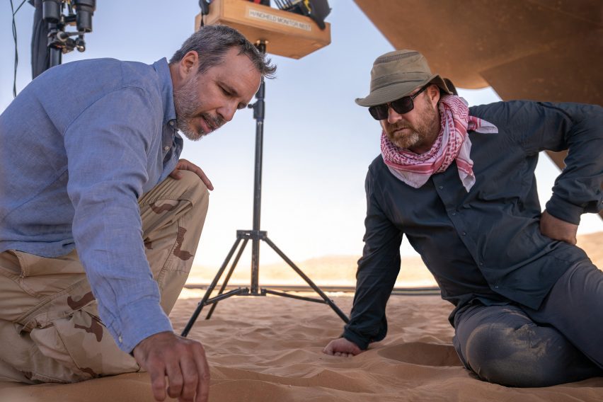 Behind-the-scenes photo of Denis Villeneuve and Patrice Vermette in discussion while sitting on the sand in an outdoor desert location