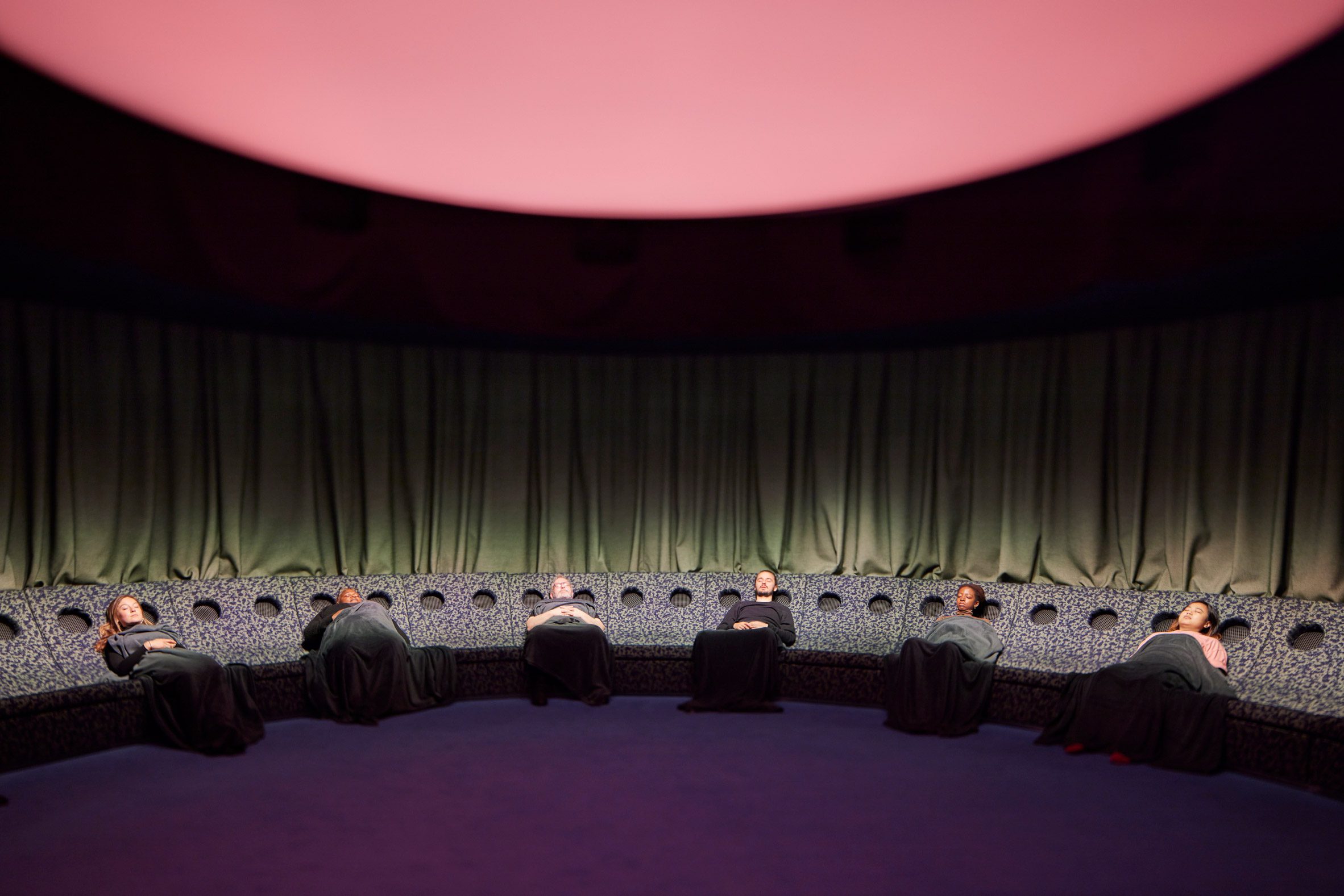 Six people laying down in a room with green curtains and a pink glowing ceiling