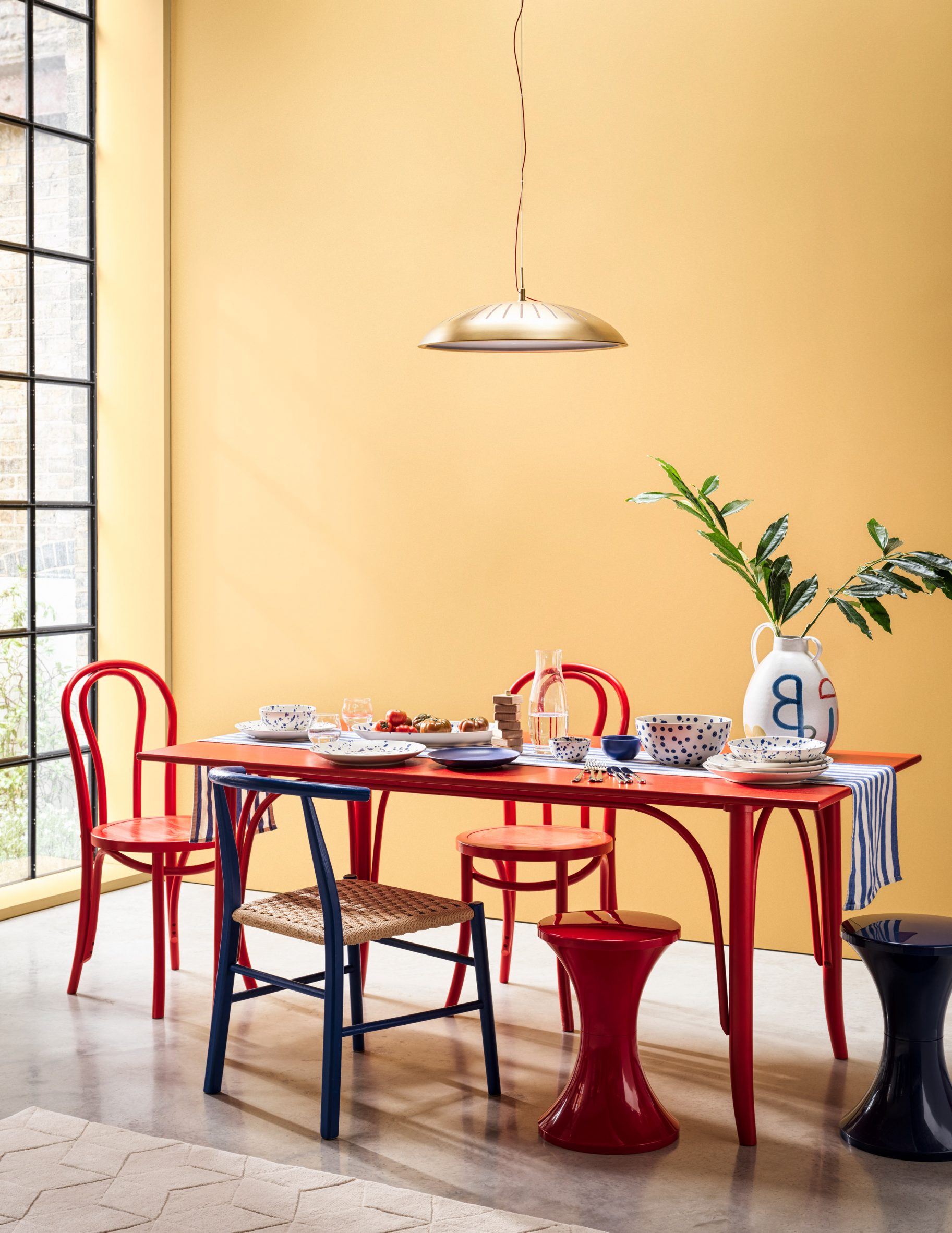 Lifestyle photo of a dinner table setting with a bright red long table and red and blue mismatched chairs, as well as tableware