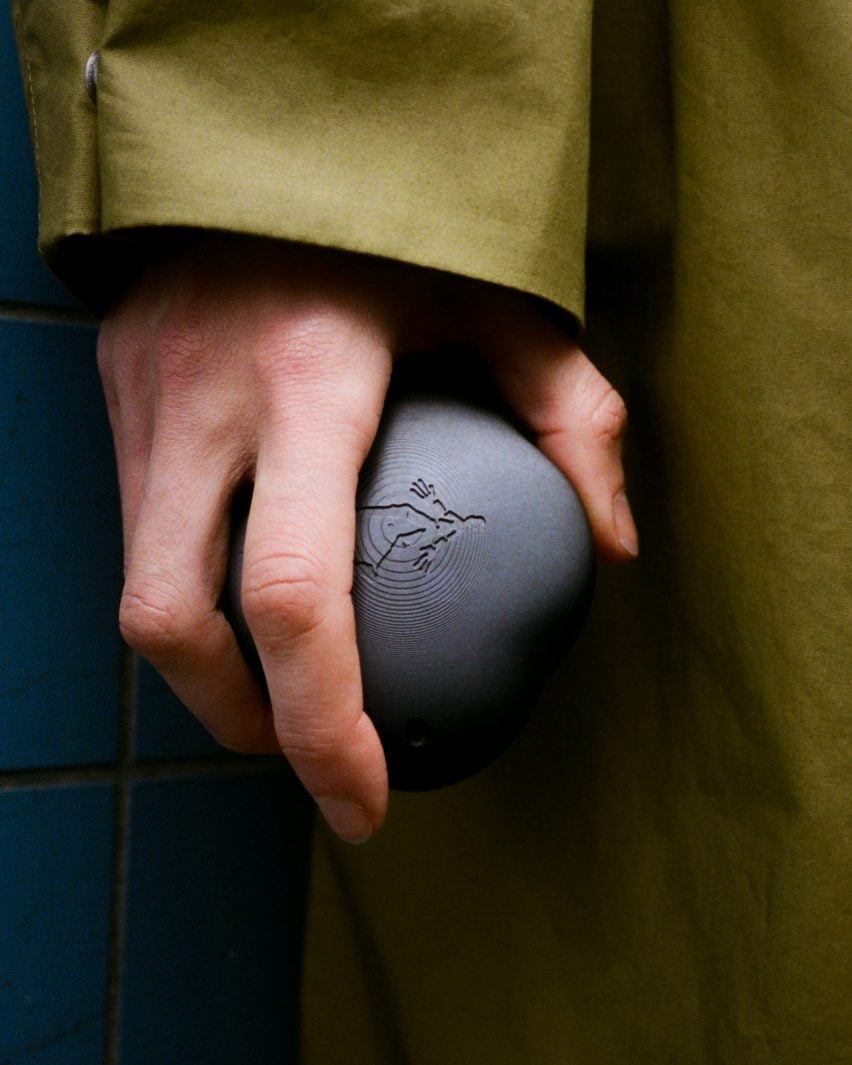 Imagen de una mano que sostiene un dispositivo con forma de roca de color gris oscuro decorado con una persona que camina 