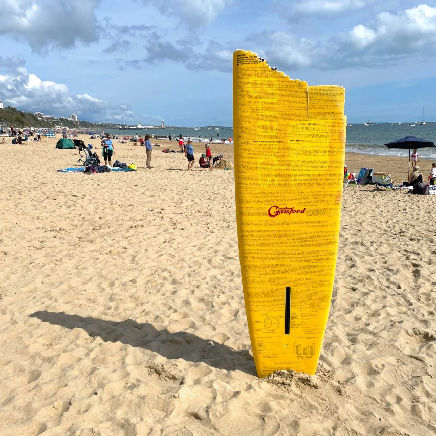 A chipped yellow surfboard with black handwriting on it placed in the sand on a beach.