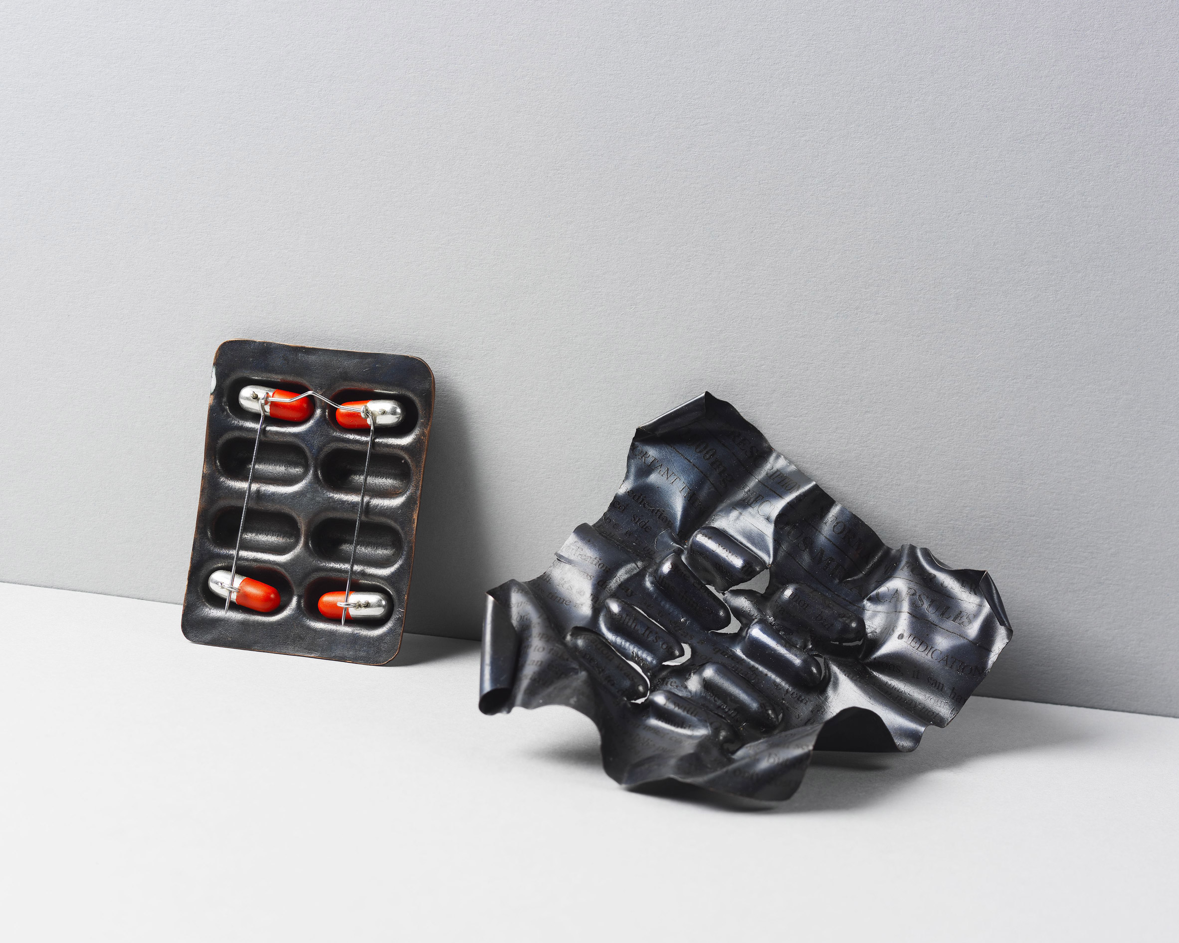 A photograph of black metal jewellery pieces in the shape of pill packets, against a white background.