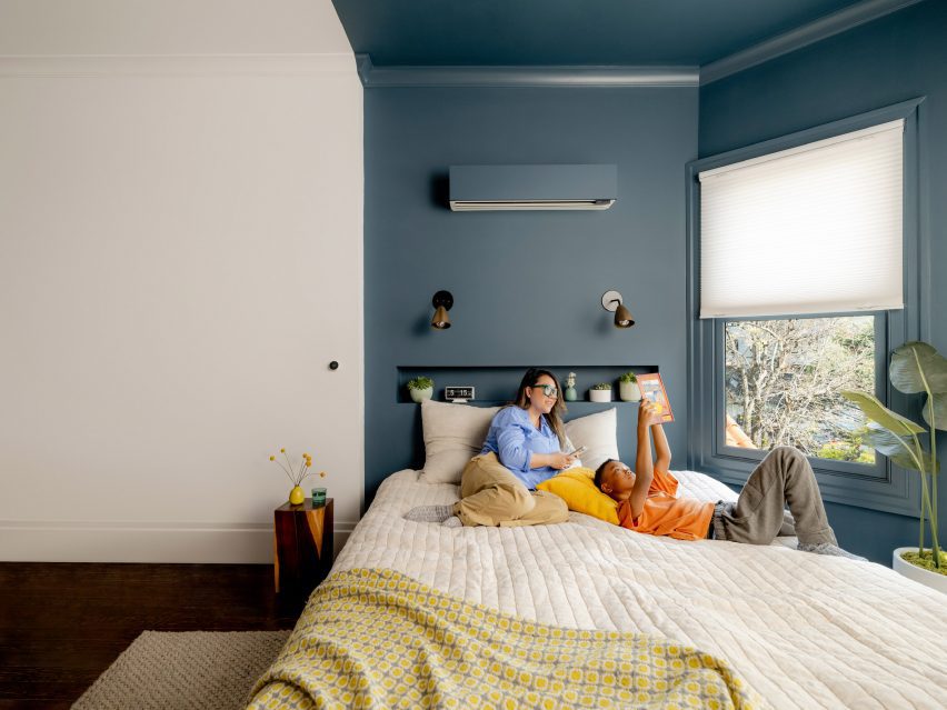 Photo of two people lounging in bed underneath a wall-mounted climate unit painted the same blue-grey as the walls