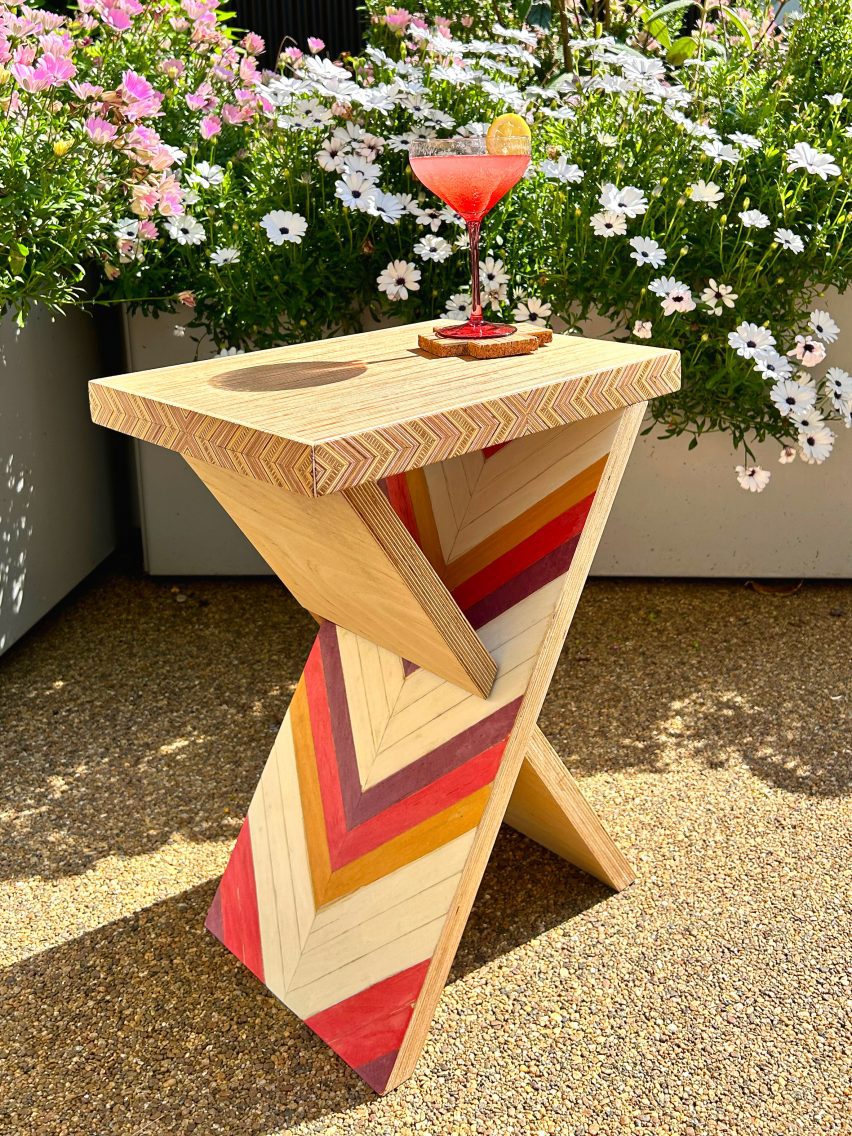 A triangular shaped wooden side table with geometric patterns in red, yellow and brown tones, placed on gravel.