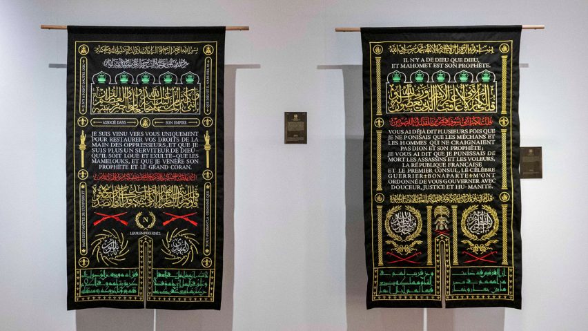 Two tapestries hanging on a white wall, black with yellow, white, green and red embroidery on them.