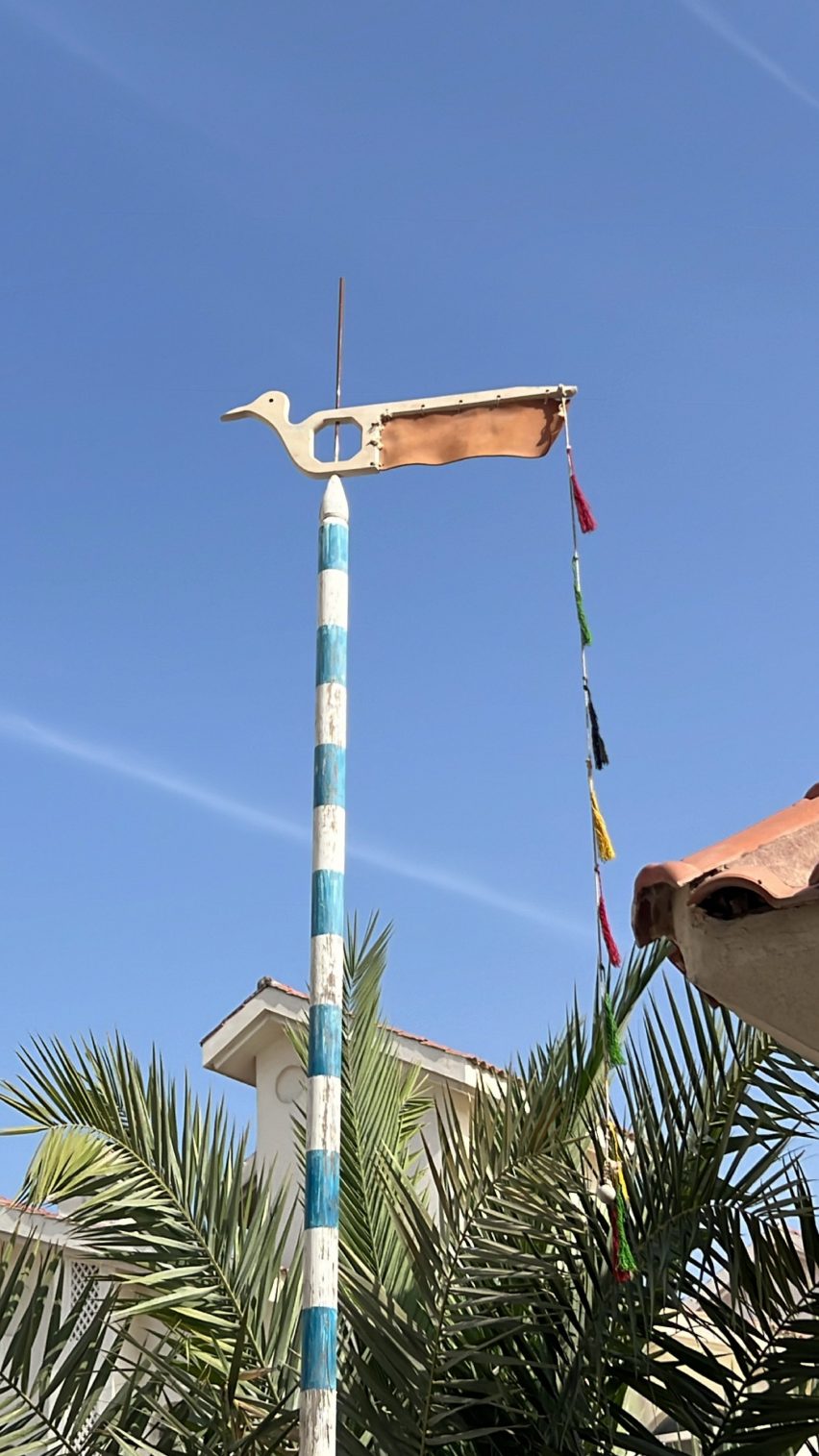 A photograph of a wooden pole in colours of blue and white stripes, with a bird figure on top. The pole is situated outside against a blue sky backdrop and a green plant.