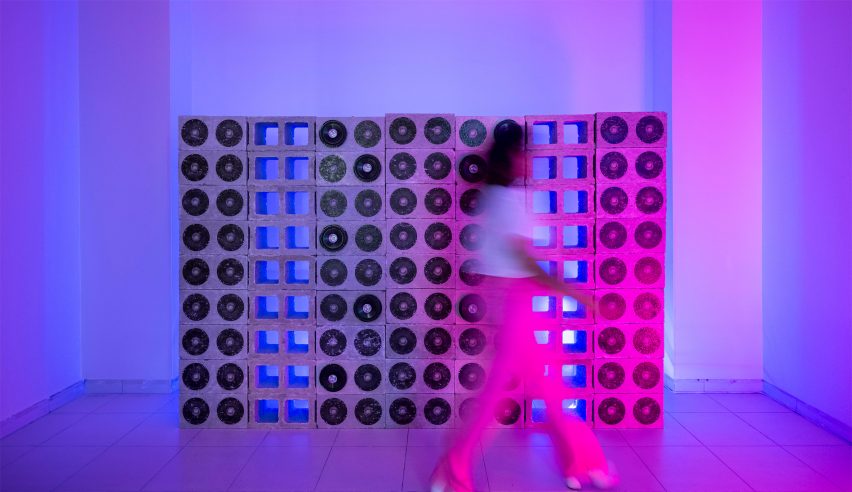 A person walking past a concrete structure with black circles on it, in a room that is lit in purple, blue and pink colours.