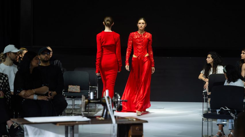 Two people walking in different directions, next to one another. Both are in red dresses and walking on a white floor with a black backdrop, with people in chairs either side of them.