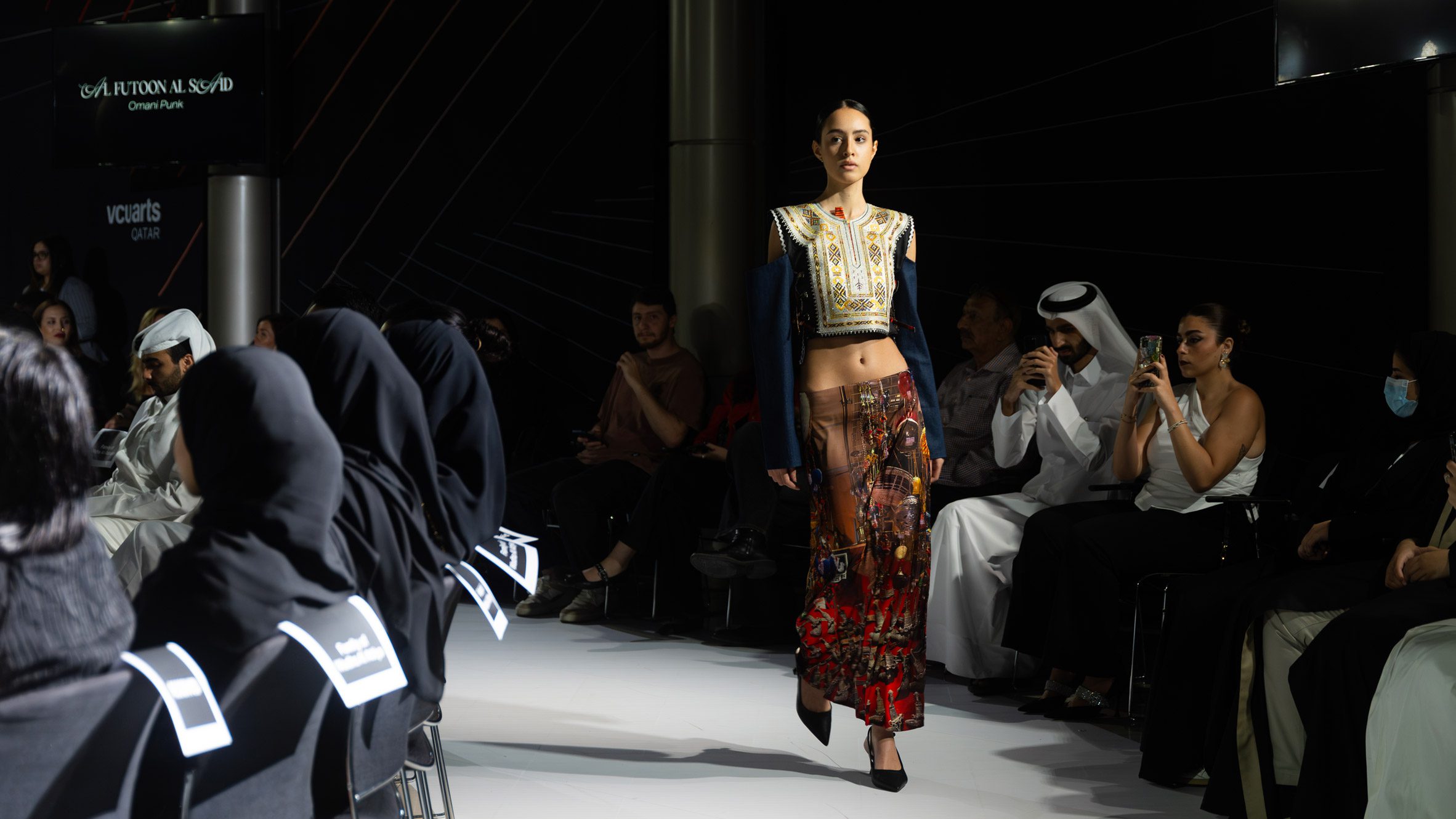 A person walking on a white runway floor with a black backdrop, wearing a red skirt and a yellow top with blue sleeves.