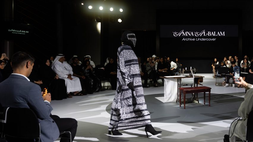 A person walking on a white floor with people in chairs sat around them, wearing black and white clothing.