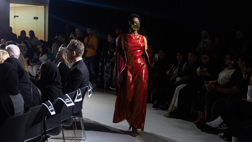 A person walking on a white runway floor with people in chairs sat around them. They are wearing a red dress and a gold garmet over their face.