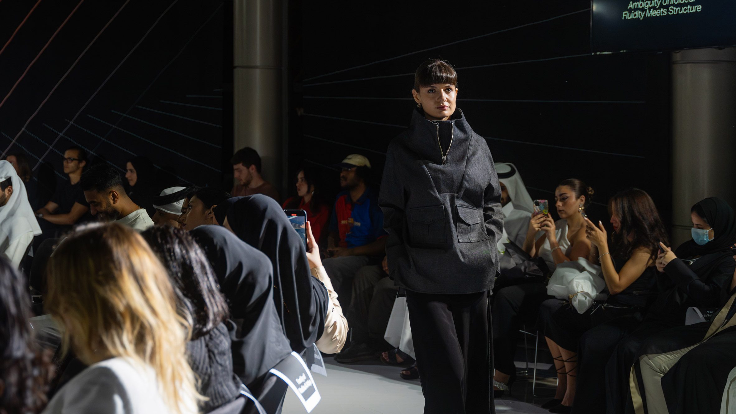 A person walking through a group of people sat on chairs, wearing black clothing.