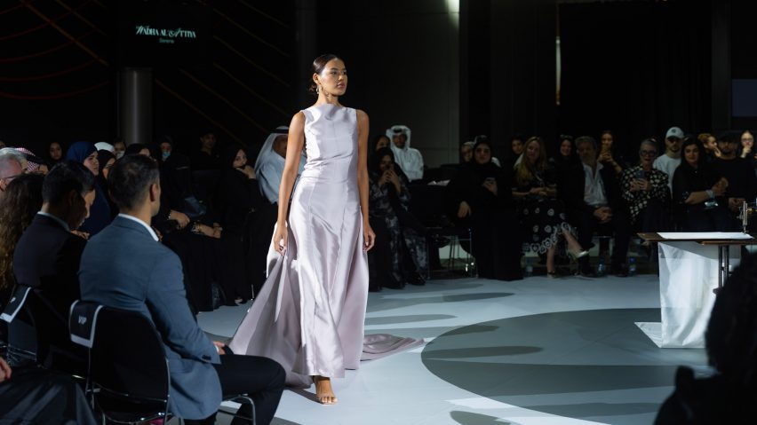 A person walking on a white runway floor with people in chairs around them. They are wearing a silver dress.