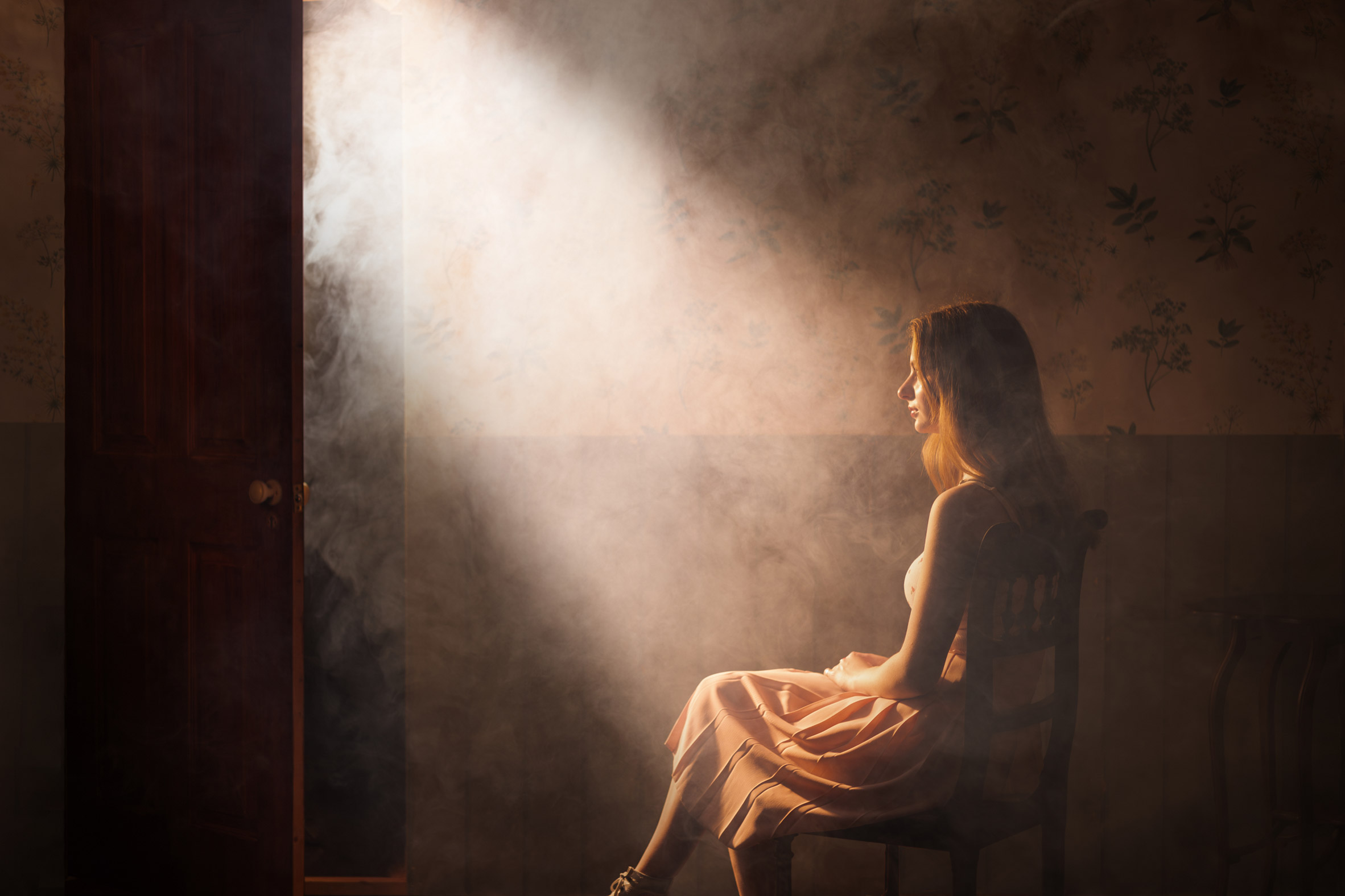 A photograph of a person from the back, sat down and looking to the side, in a dark setting with smoke around them.