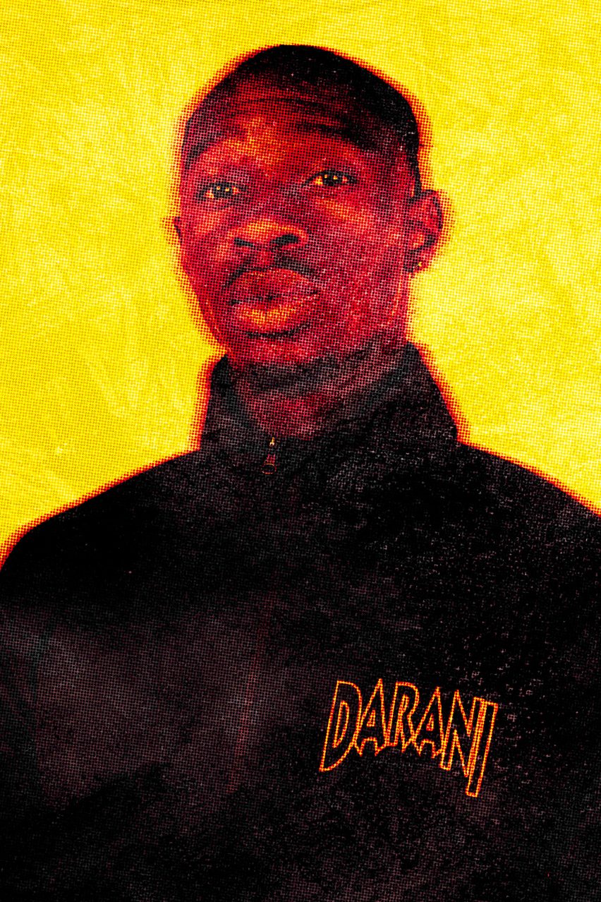 A photograph of a person looking directly at the camera, wearing a piece of black clothing with the word 'darani' written on it in yellow writing. 