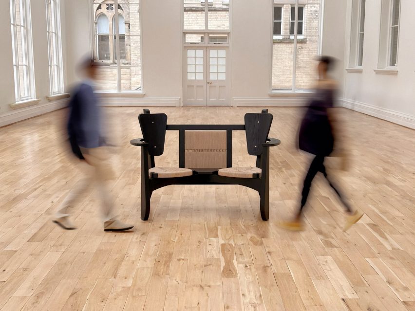 A photograph of a dark brown chair on a wooden floorboard, with blurred people passing by it.