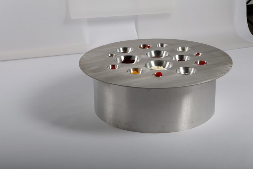 A photograph of a circular metal table, silver in colour, against a white background, with various holes on the tabletop which contain different foods.