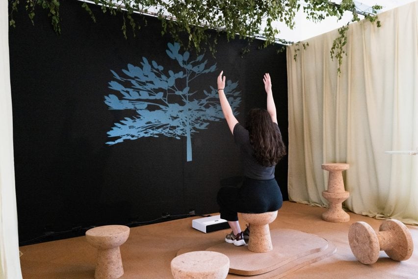 A photograph of a person sitting on a brown stool with their arms in the air, facing a large image of a blue plant against a black backdrop.