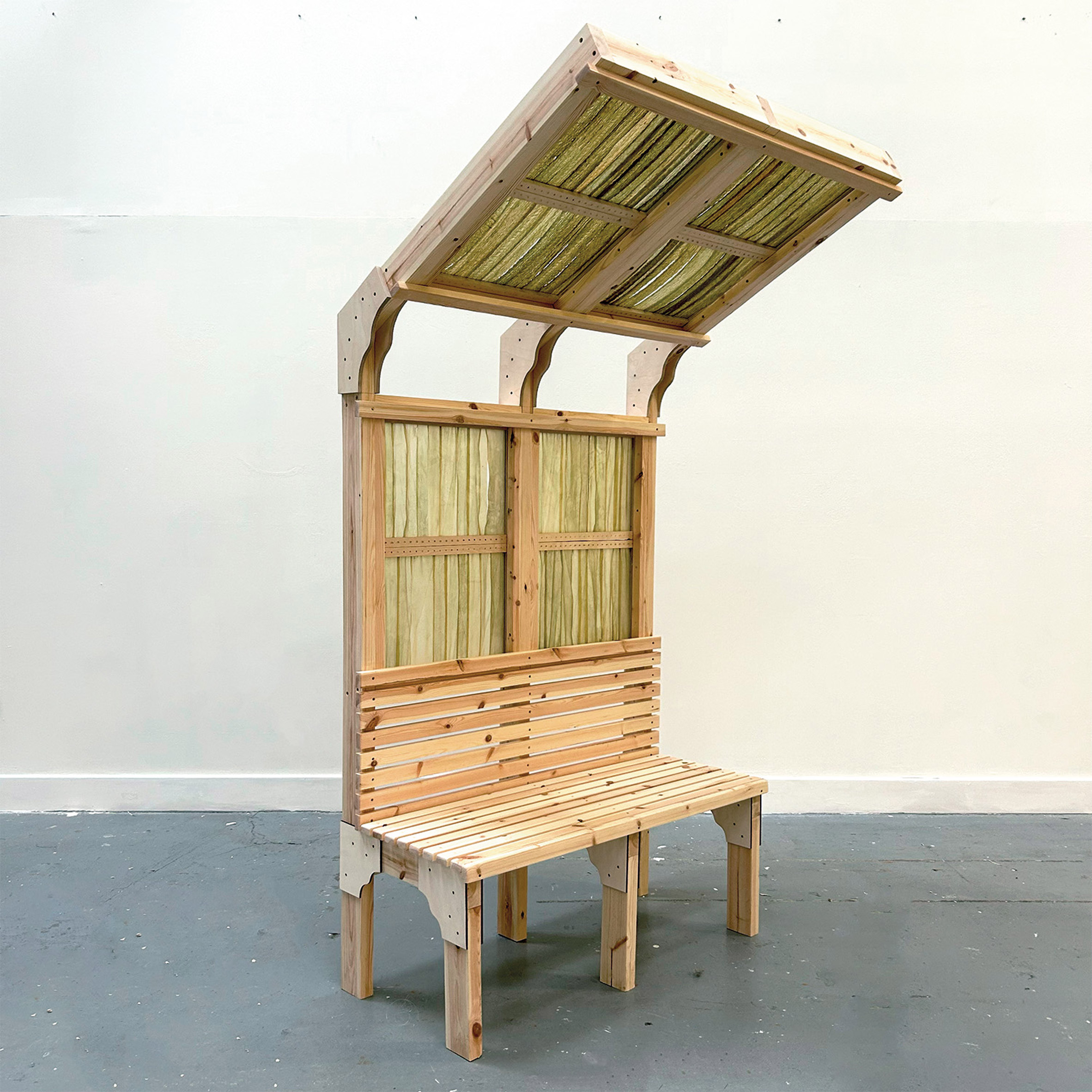 A photograph of a wooden seating structure with an angled shelter over it, made from wood and green seaweed. 