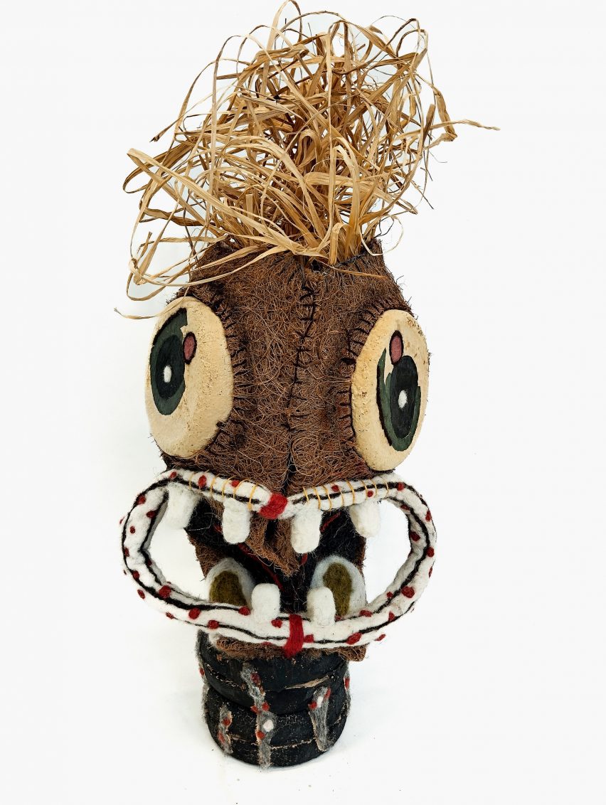 A photograph of a textiles object, which resembles a character, in tones of brown, green, white, red and black, against a white background.