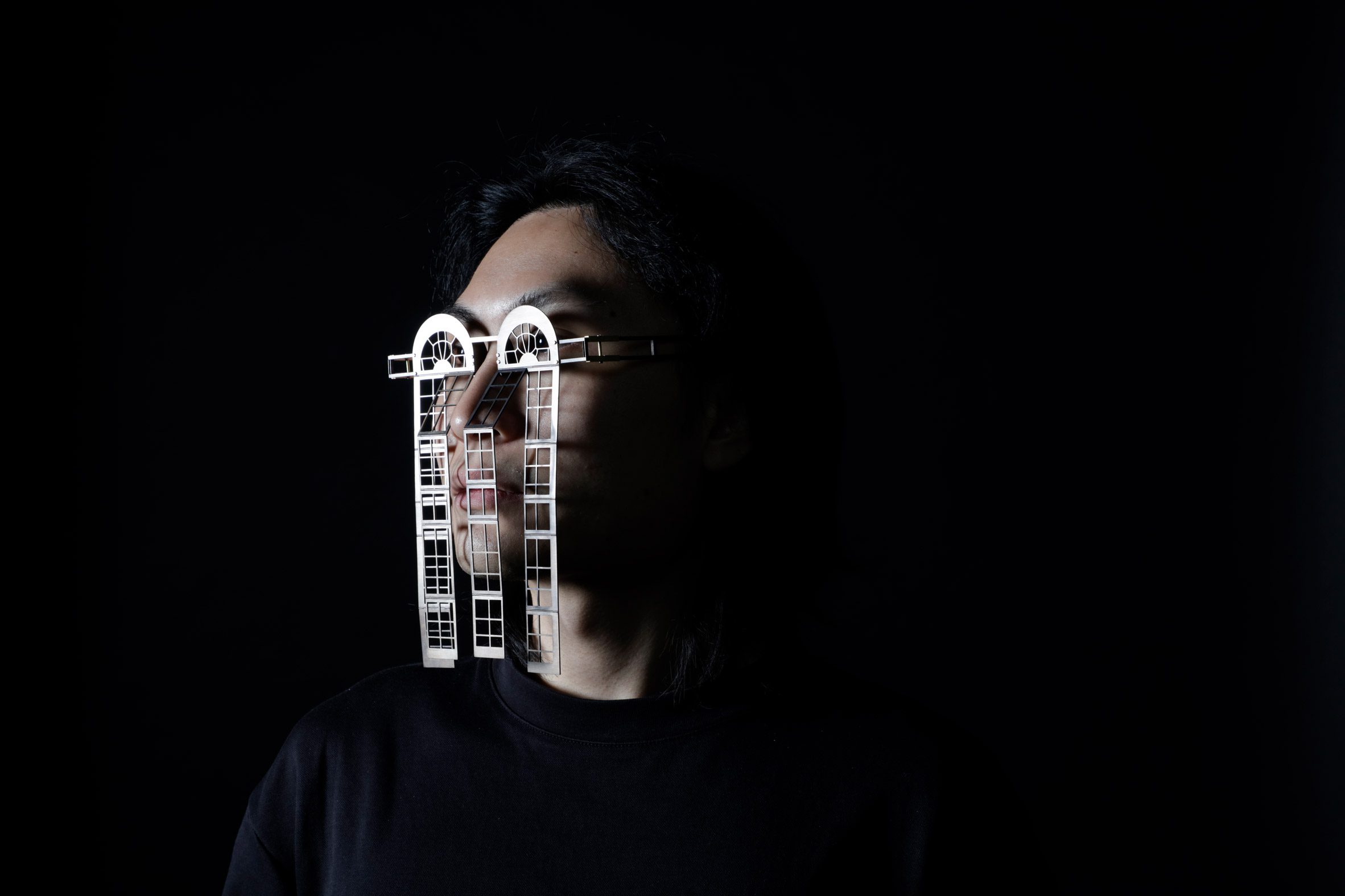 A photograph of a person's head, against a black background, wearing silver metal glasses that replicate architectural details.