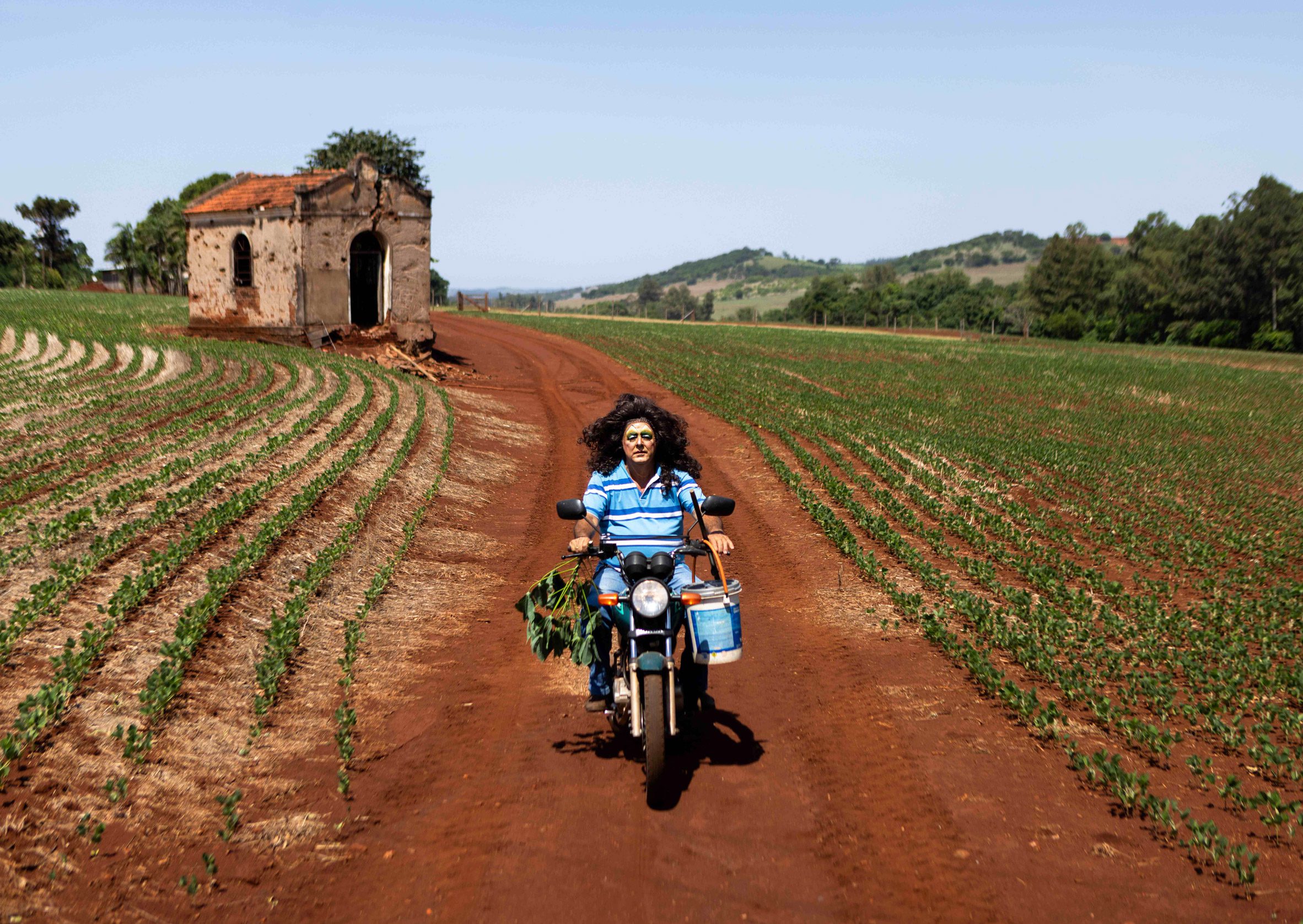 A photograph of a person in a blue shirt riding a motorbike through a field. There is a small stone hut behind them and a blue sky.