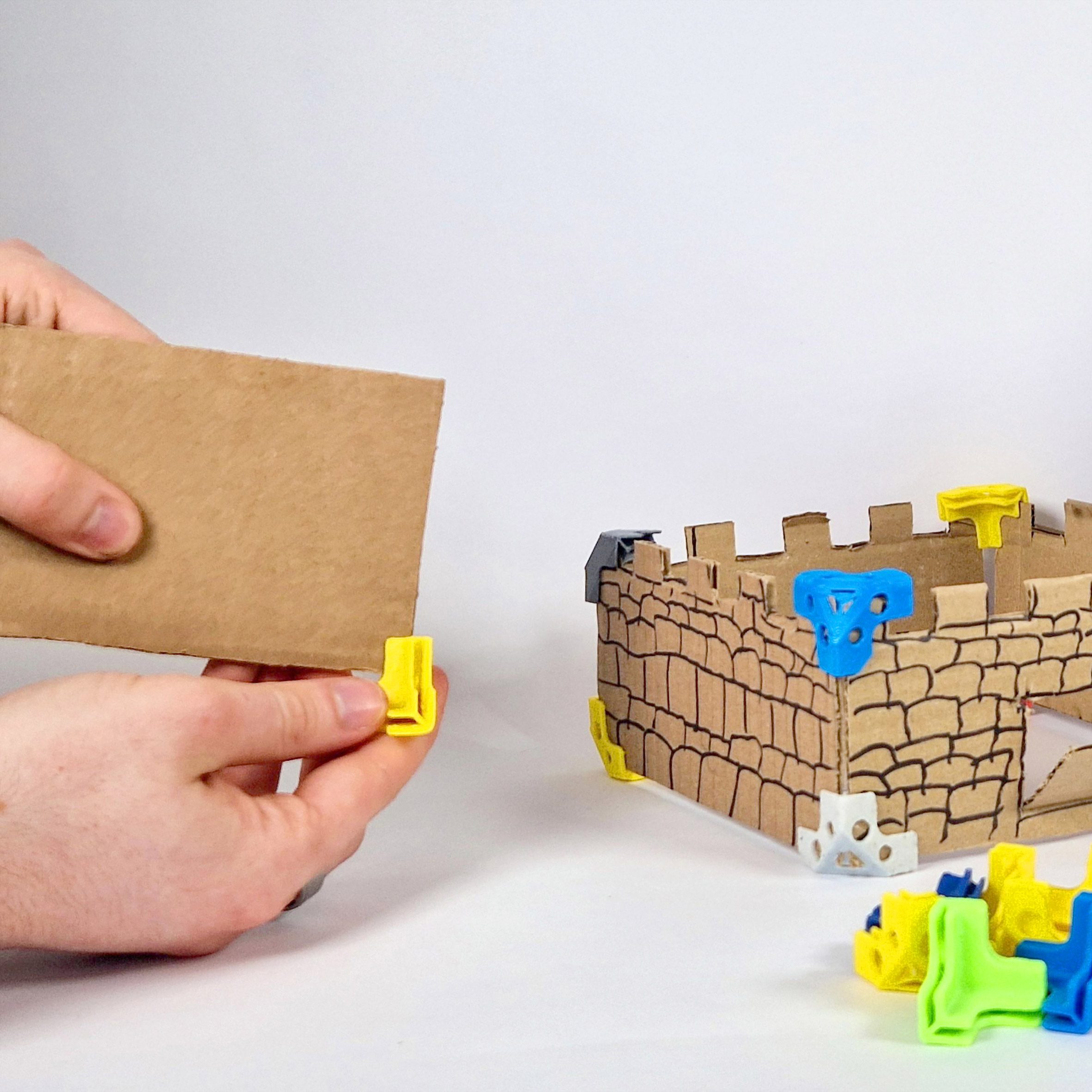 A photograph of a person handling a brown rectangular material, next to 3D rectangular cardboard model, with blue, green and yellow fastenings on its corners.