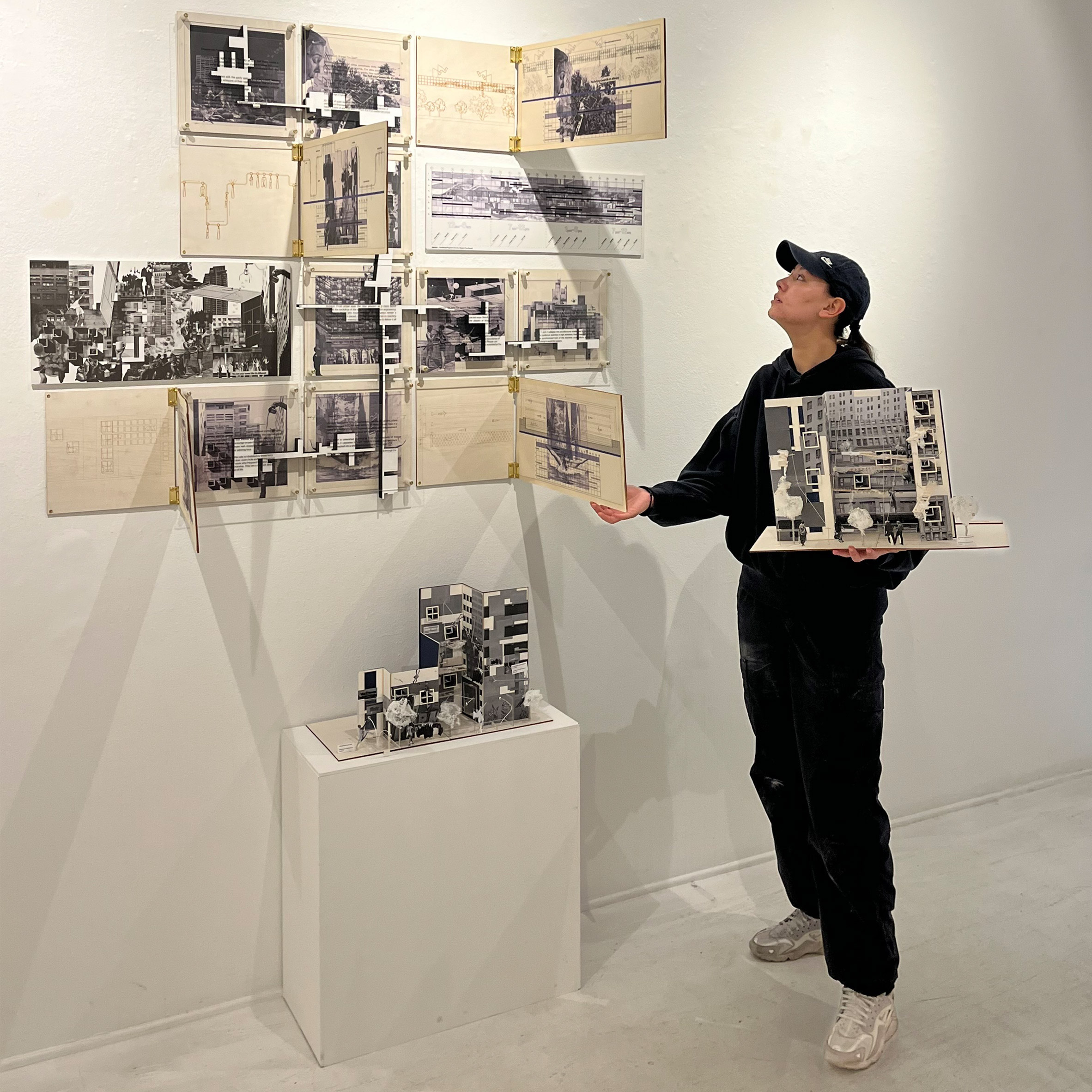 A photograph of a person holding an architectural model, next to an installation on a white wall behind them.