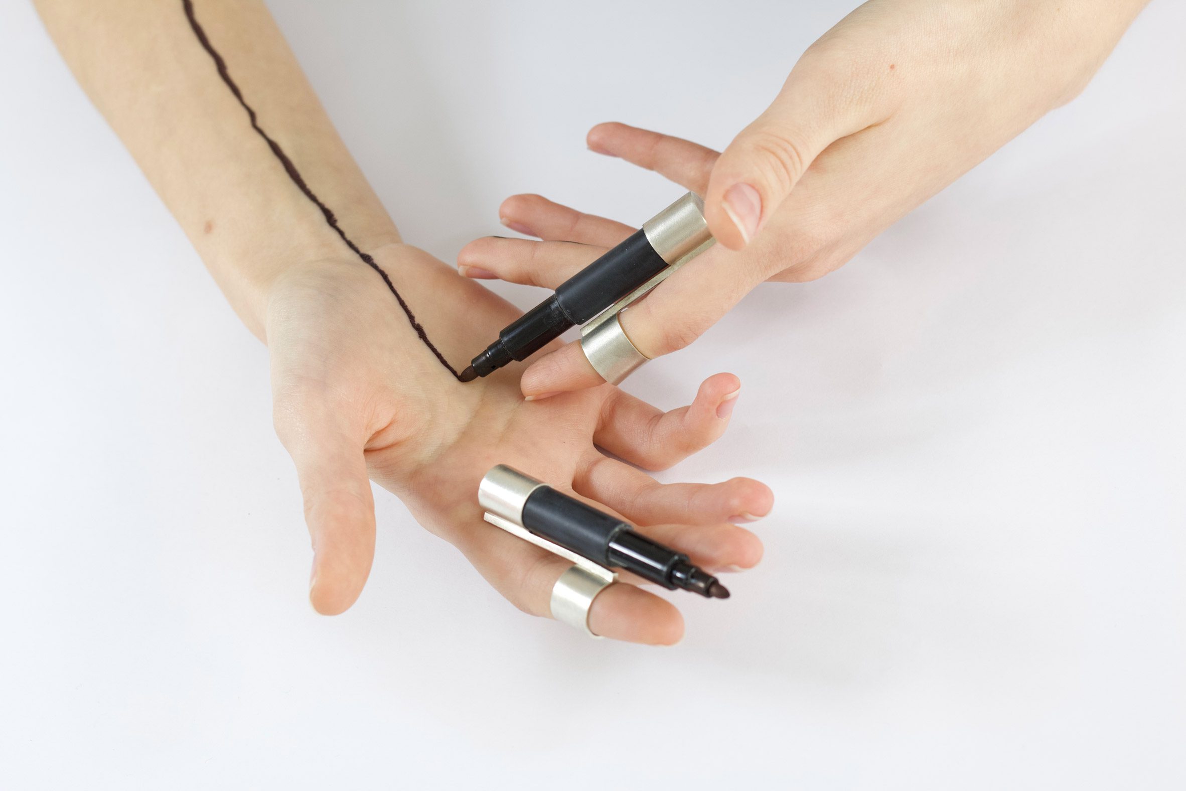 A photograph of a person's hands with a ring on each one, with black pens attached to them.