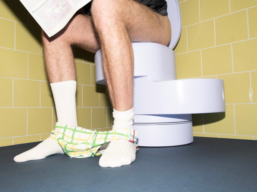 A photograph of a person's legs sitting on a toilet, with white socks on and underwear around their ankles. The walls are yellow and the ground is grey.