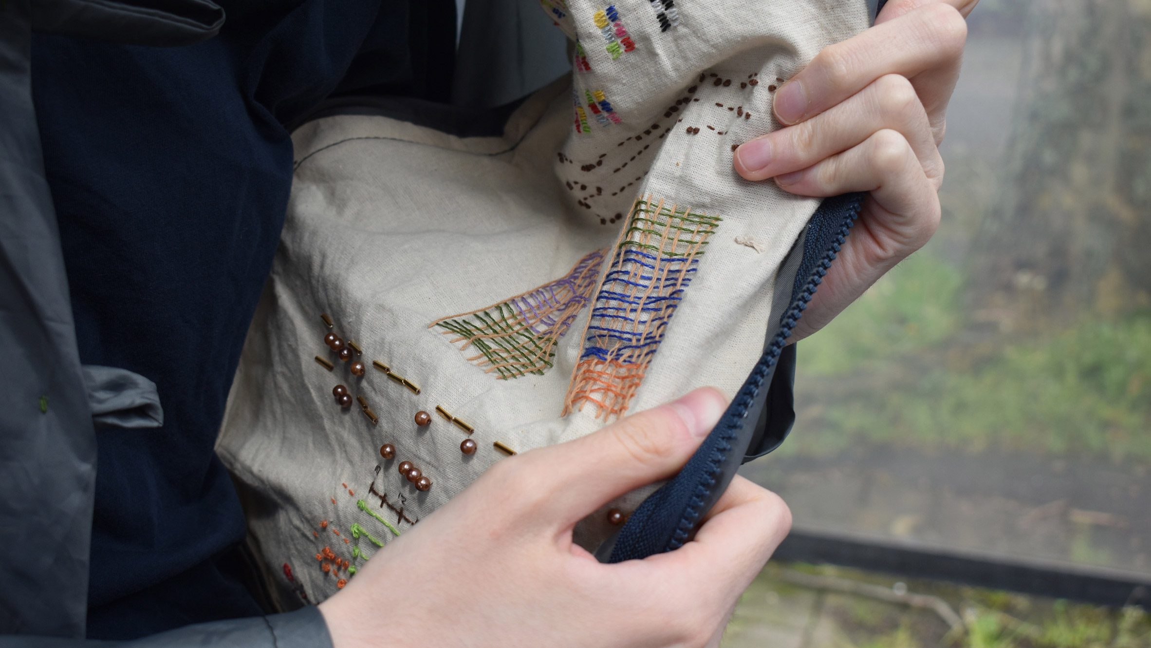 A photograph of a person holding a beige fabric with multicoloured stitching on it.