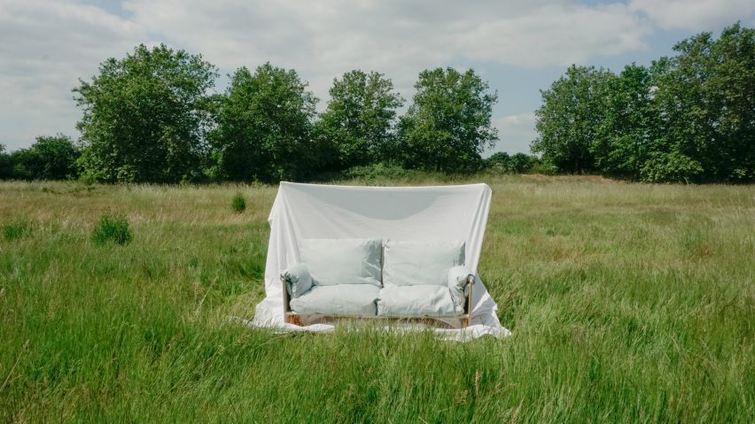 A photograph of a sofa covered in fabric, with a sheet of fabric held up behind it, placed in a green field.