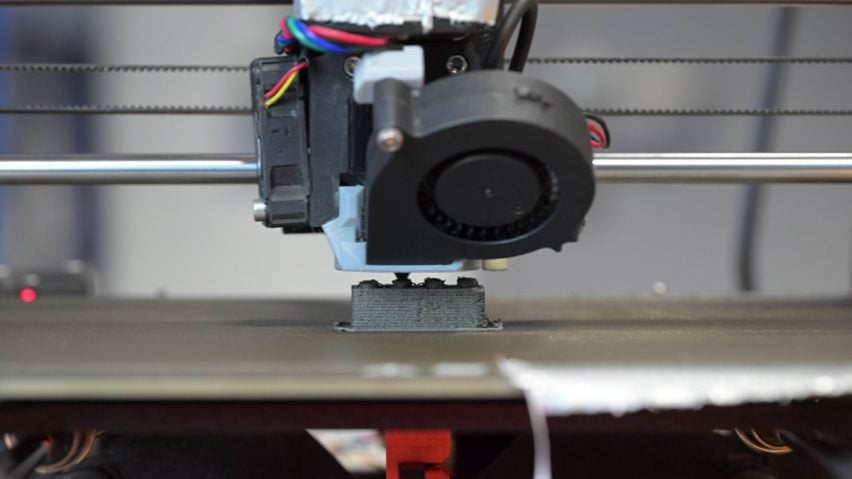Close-up photo of a 3D printer manufacturing a small, grey, Lego-like block