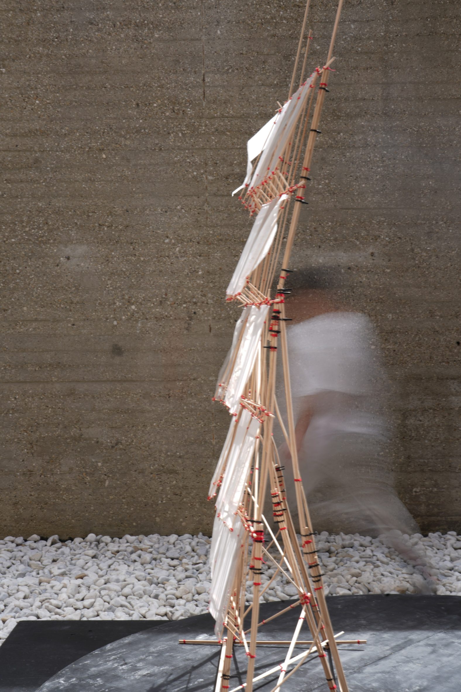 A photograph of a geometric structure composed of brown sticks and white rectangular sheets, against a brown background with a blurred figure passing.