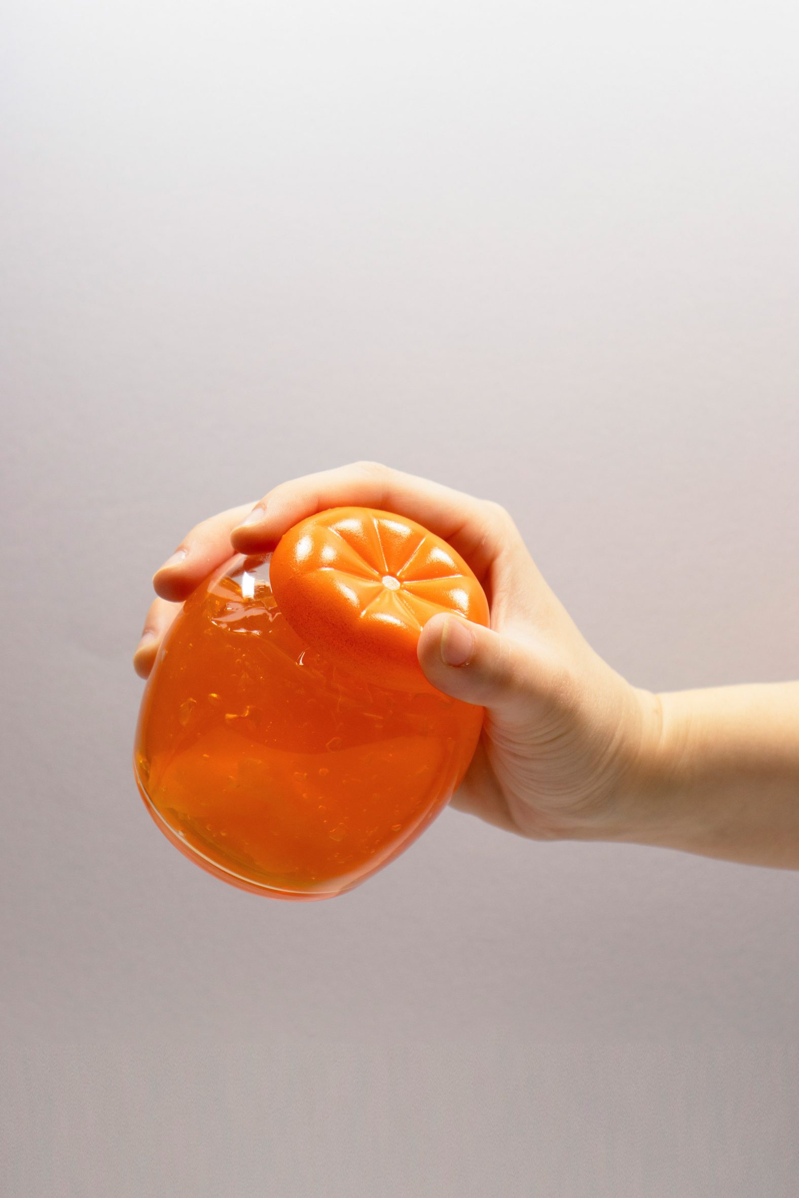 A photograph of a person's hand holding an orange coloured circular object against a white background.