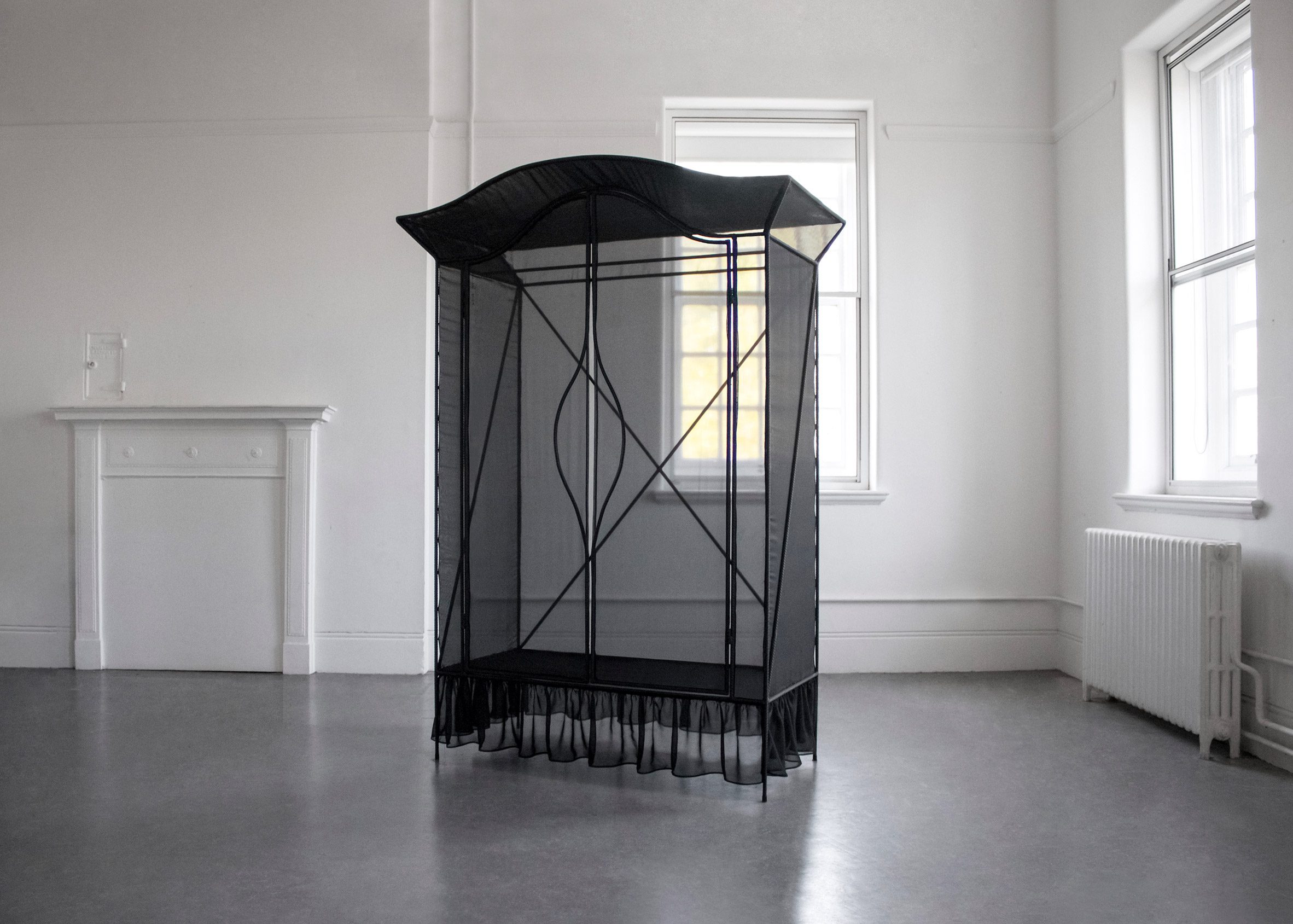 A photograph of a black wardrobe frame covered in black netting in a white room