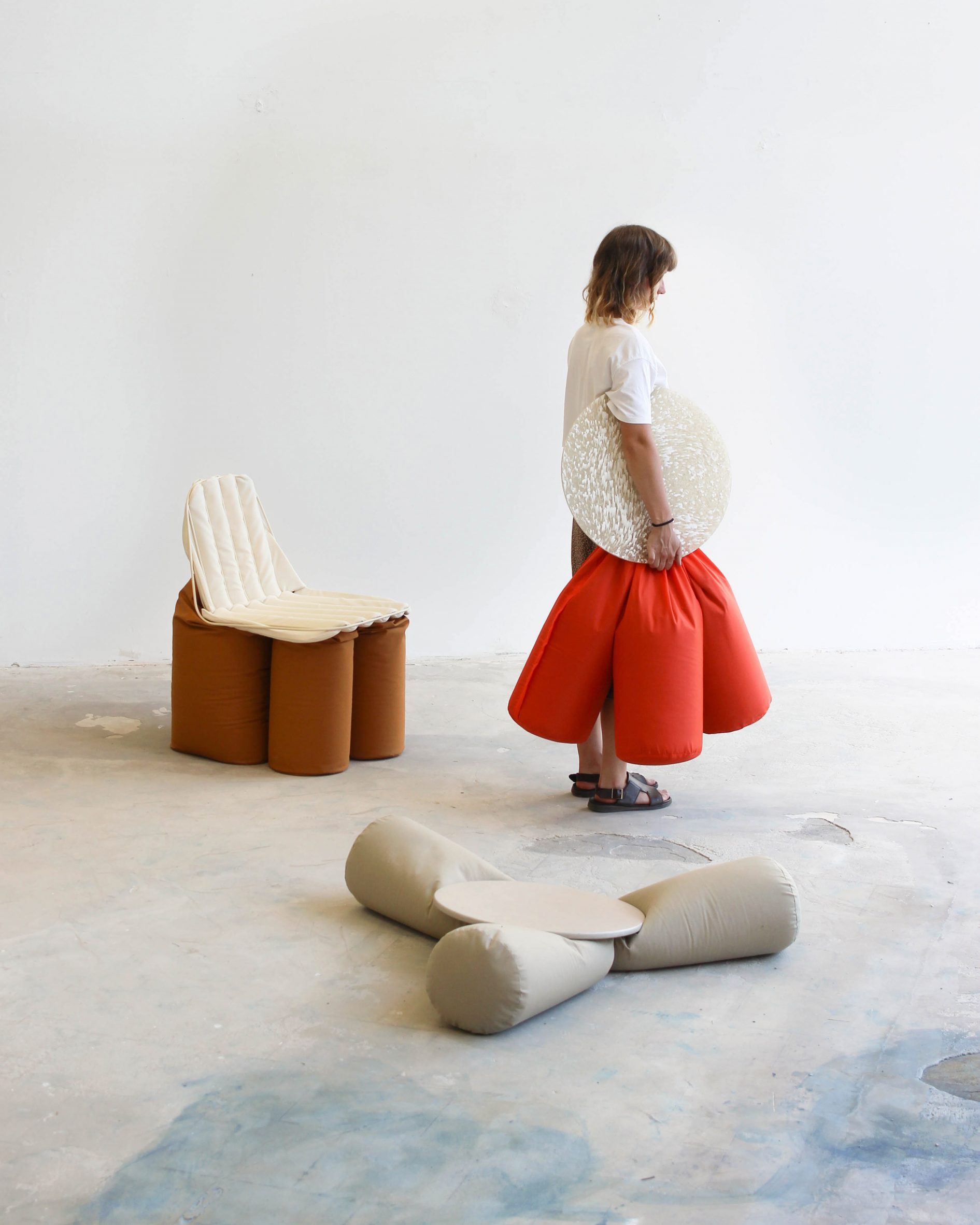 A photograph of a person, facing to the side, holding a seat featuring an orange cushion and a beige circular surface. There are two other seats surrounding them in tones of grey and brown. 