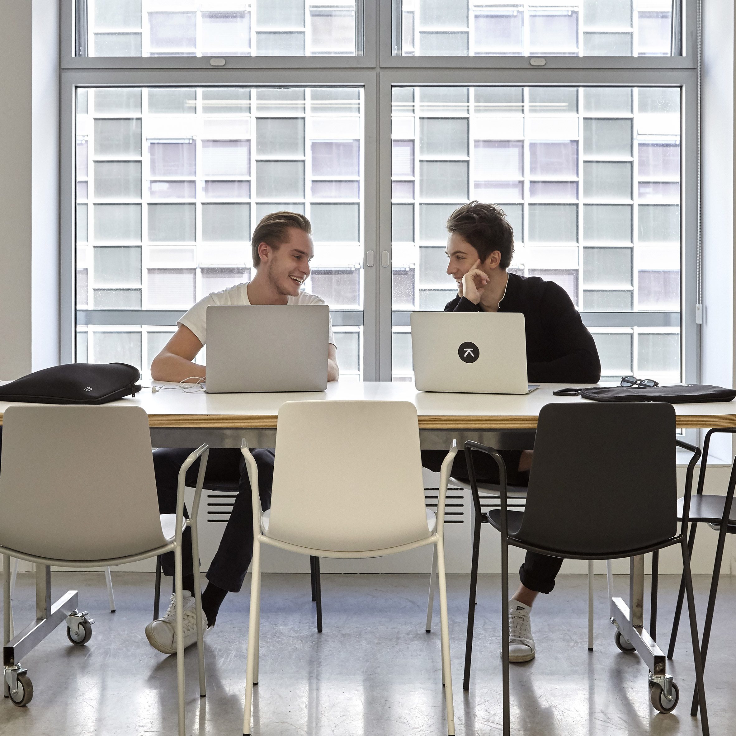 Two students sat at a desk conversing with their laptops open.