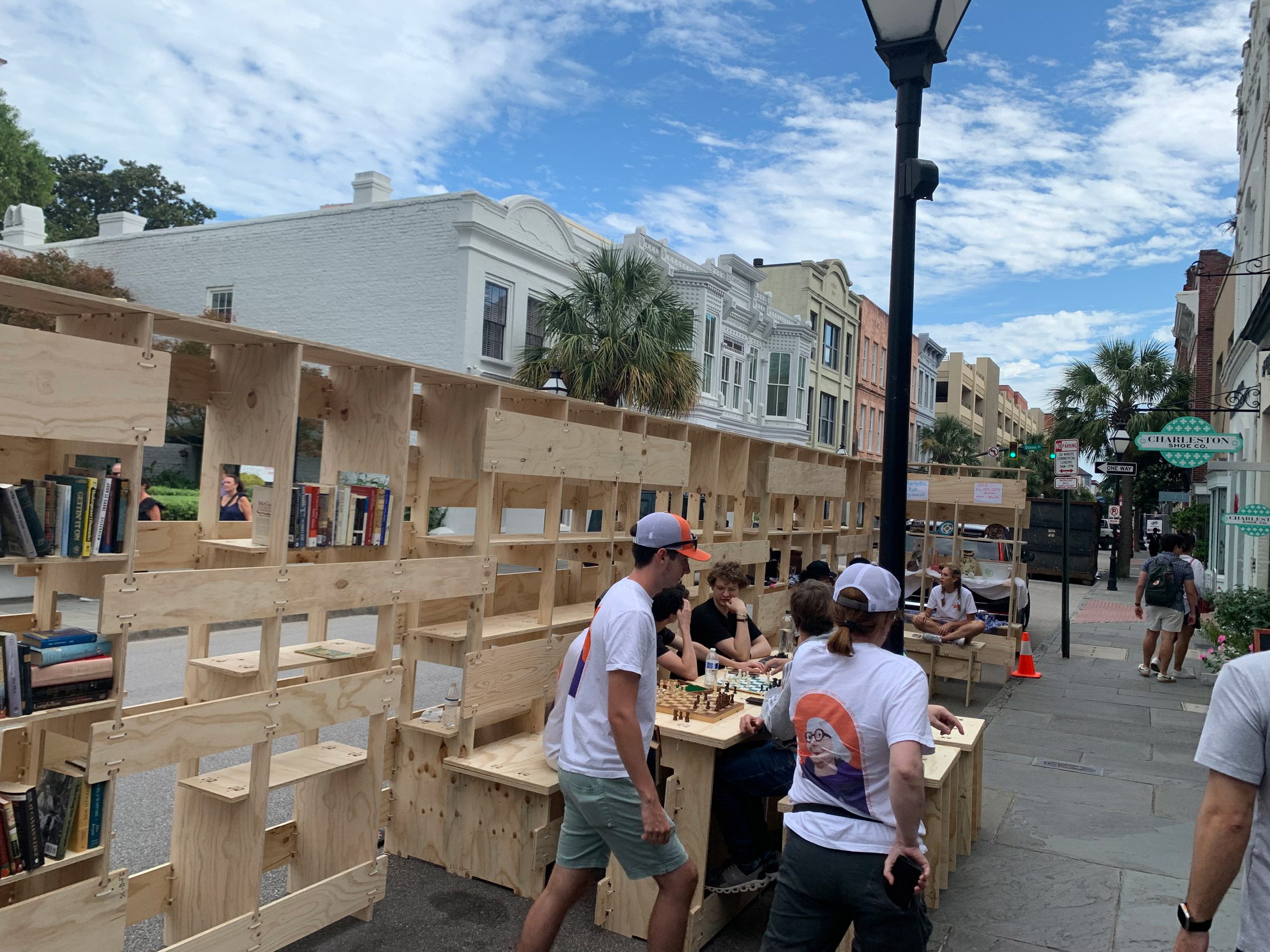 A photograph of a wooden structure in a street with, people passing by and others sat at it playing chess.