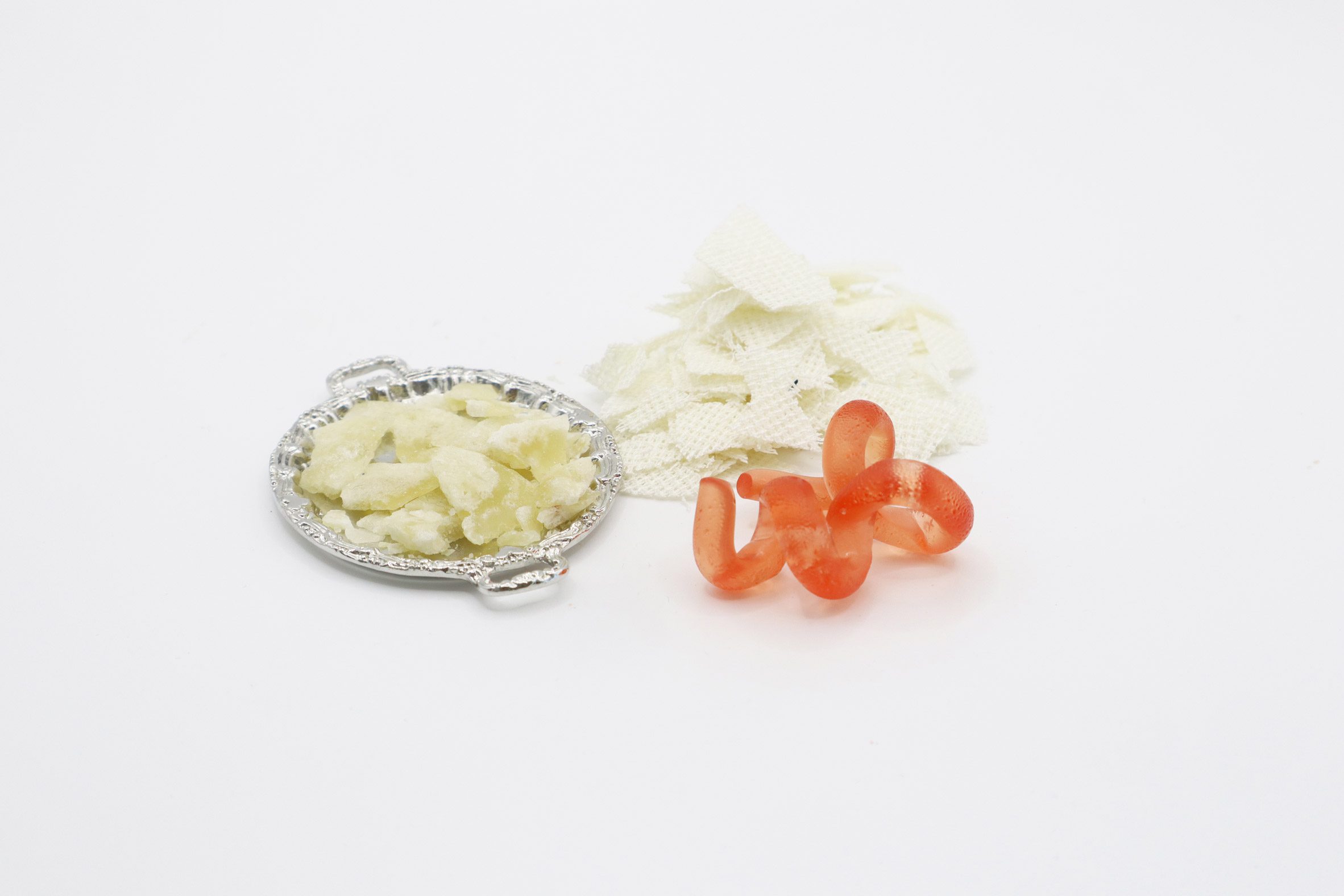 Photo of off-white fabric scraps, a similarly coloured mystery material on a small plate and a bright orange twisted Snake-like gummy candy