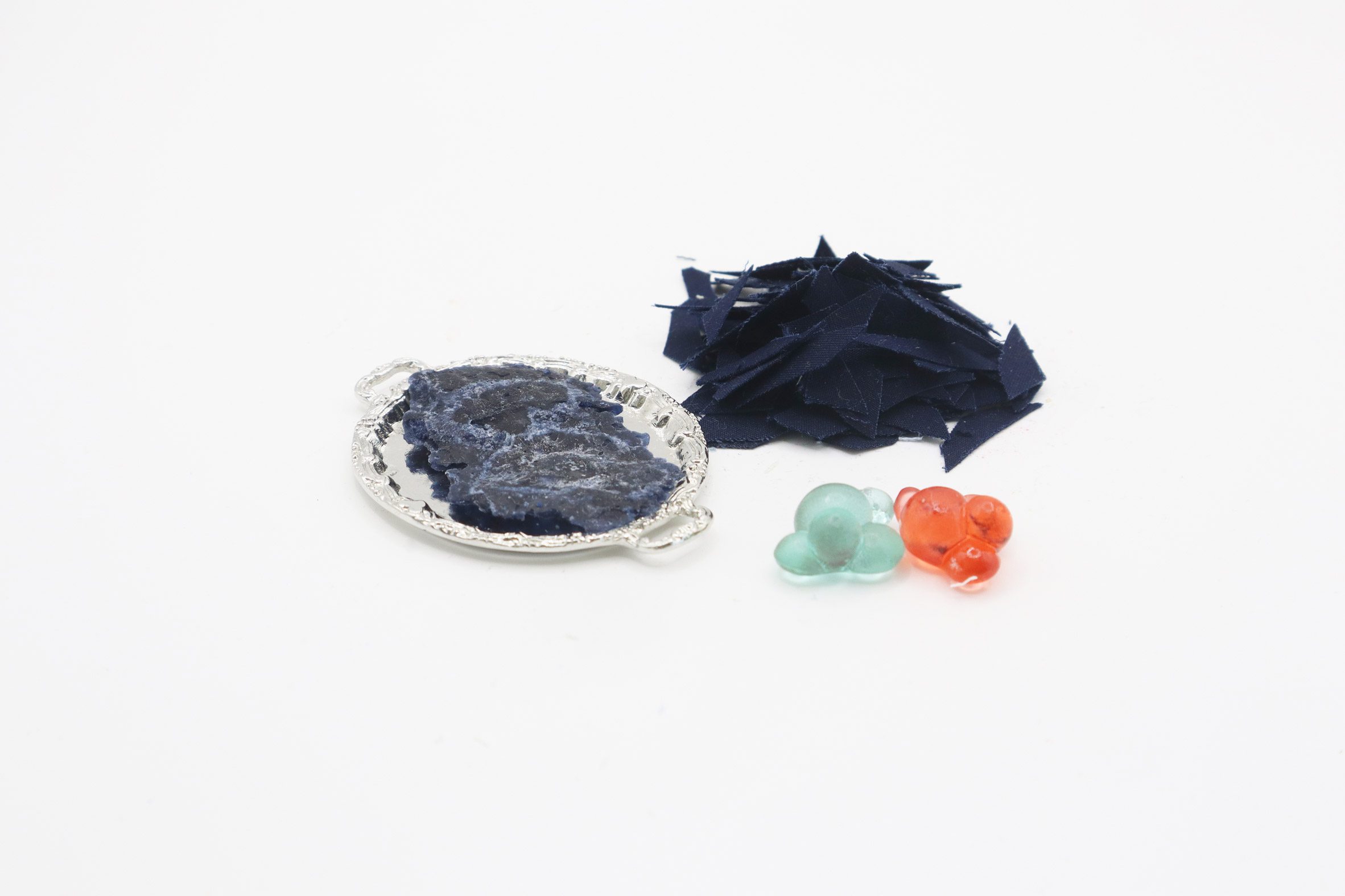 Photo of scraps of navy blue fabric beside a small platter of partly broken down fabric and two multi-coloured gummy sweets in abstract shapes