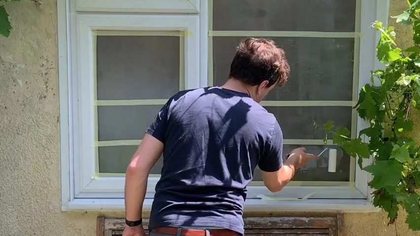 Video still of a man painting the outside of the windows on a house with a translucent, paint-like substance