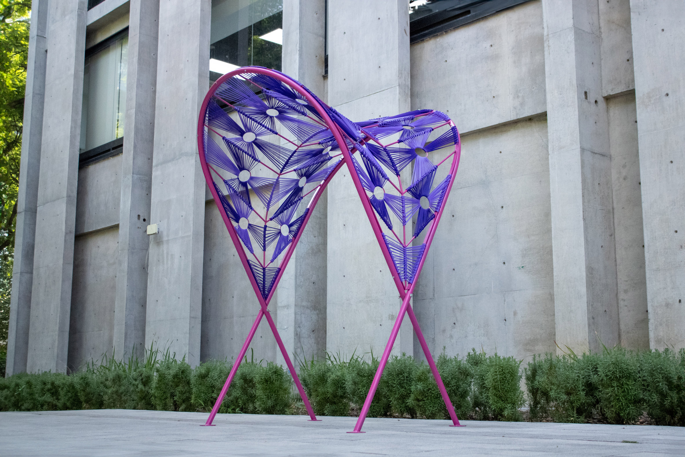 A photograph of an outdoor installation in tones of purple and pink against a grey concrete wall. 